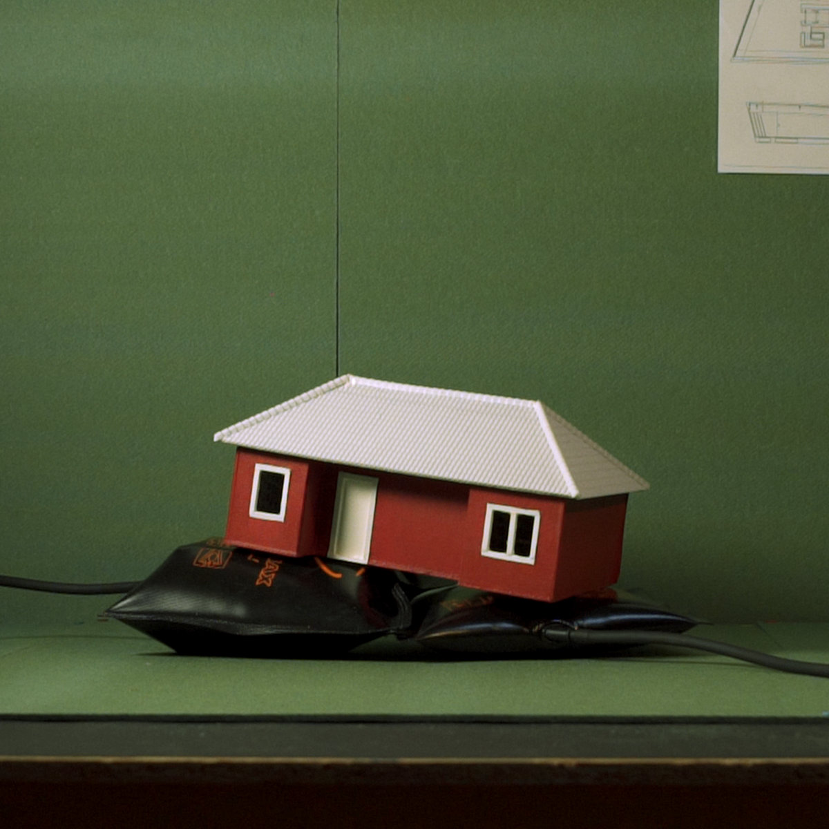 Photo of a scaled down 3D mockup of a red house with white roof, on top of what appears to be a blood pressure’s air bag. The background is green like moss.